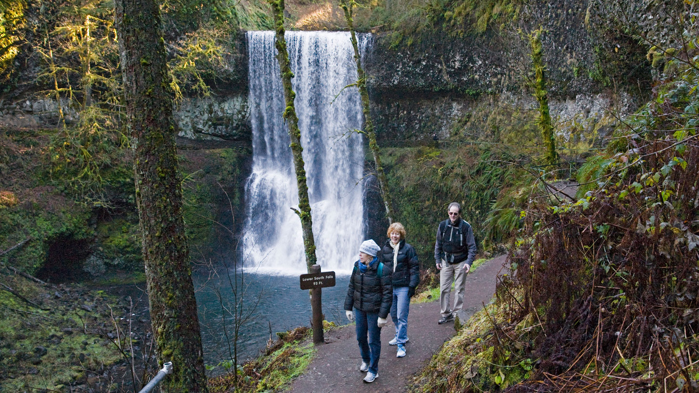 People walk along a trail with a waterfall in the background.
