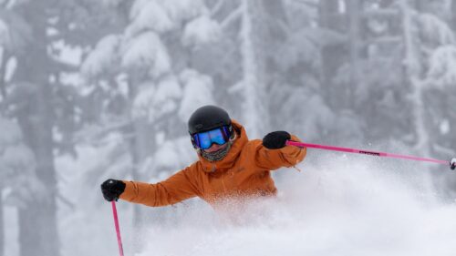 A skier in an orange jacket turns in deep powder.
