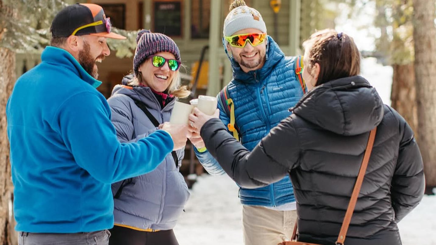 A group of four people outdoors in winter gear cheer their drink glasses.
