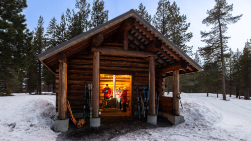 A view from outside a wooden cabin in the snow. Inside, you can see several people huddled around a wood stove.