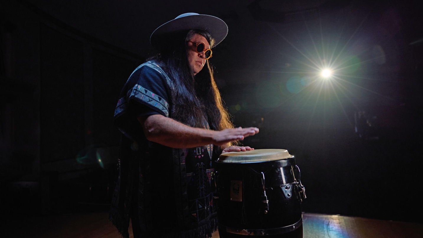 Man with long hair, hat and sunglasses plays a drum in a dark room