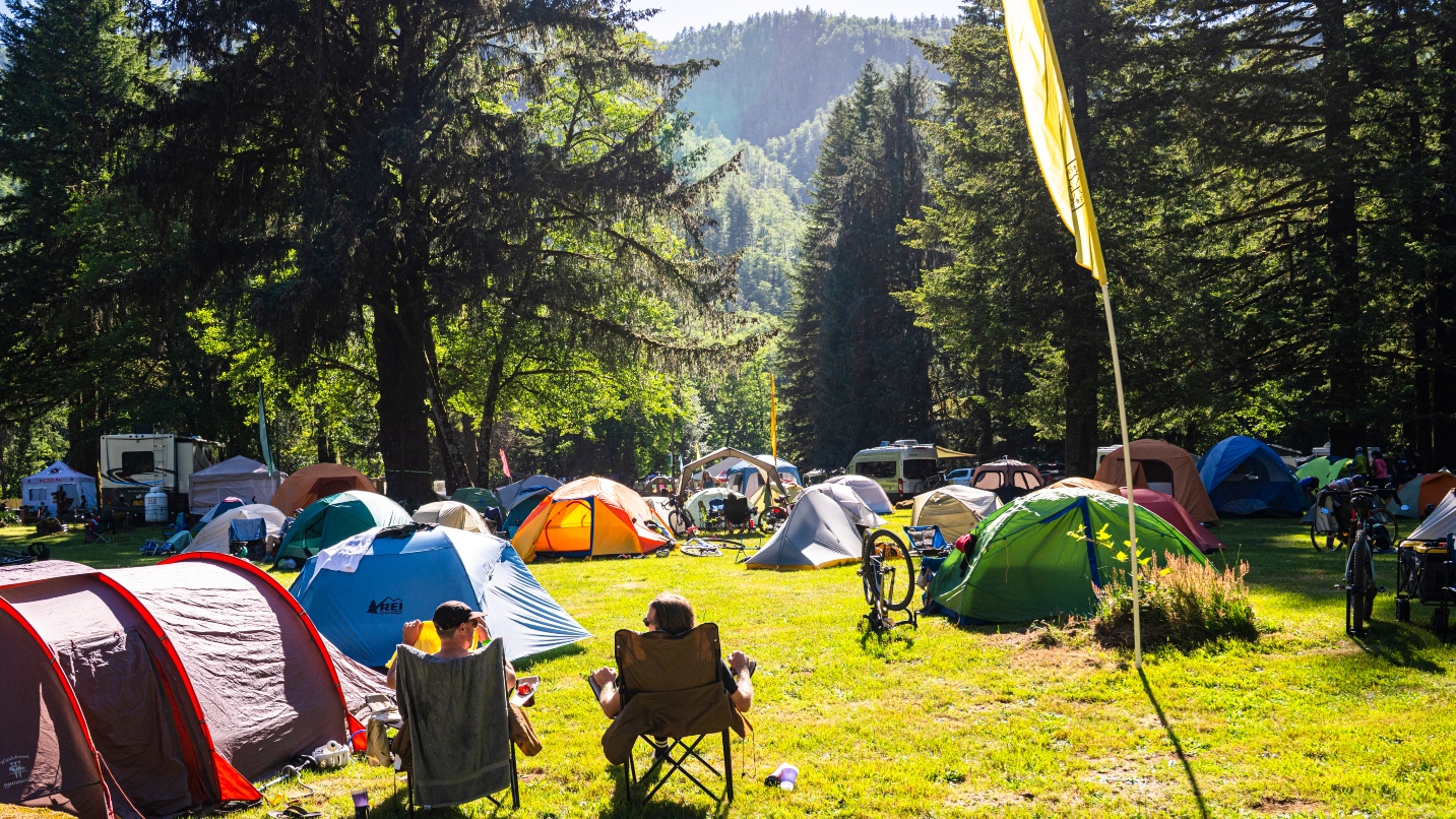 Two people sit on camp chairs with many tents on grass in front of them in a forest