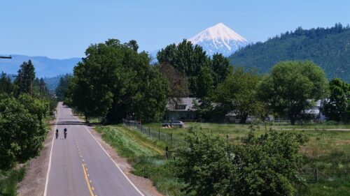 Two people ride bikes on a straight road with tree and a snow-capped mountain in background