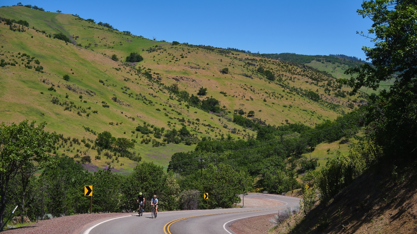 Two people ride bikes on a winding road against a green mountain backdrop
