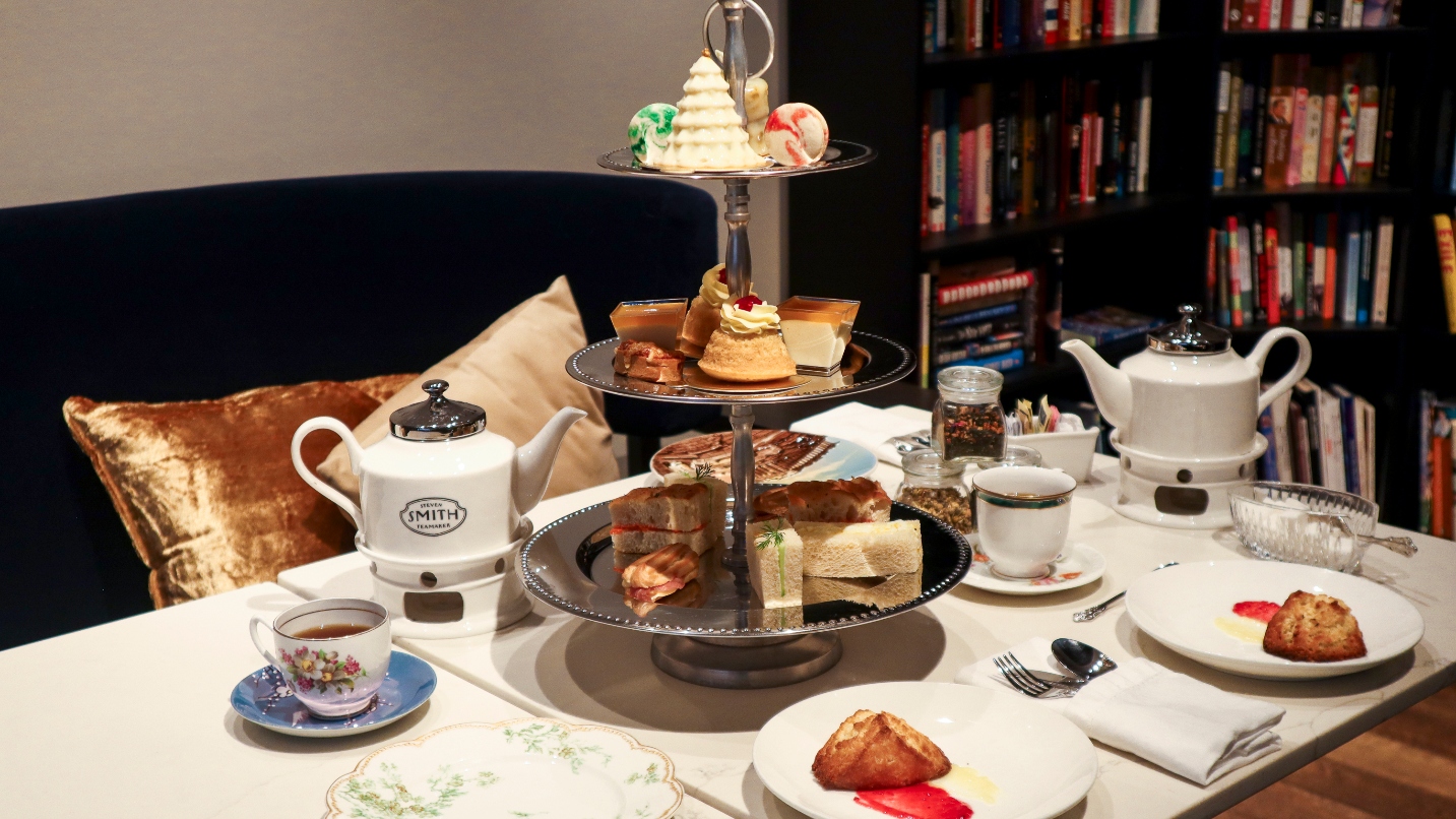 Silver tiered tray and plates of treats with two white teapots on table and bookcase in background