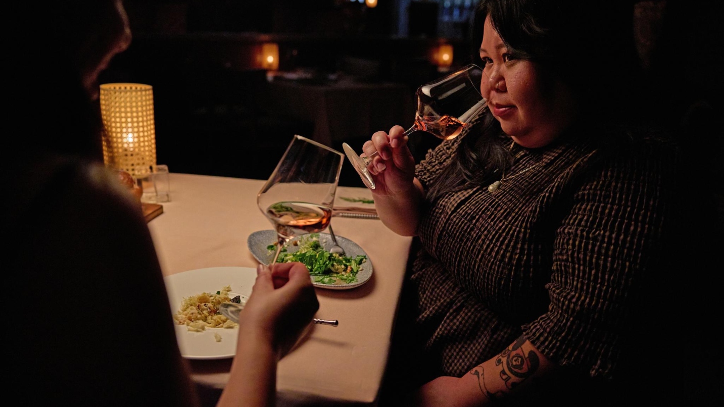 A woman holds a wine glass to her nose as she sits at a dark restaurant with a dining companion