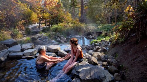 Two women sit in a hot spring with stones and forest around