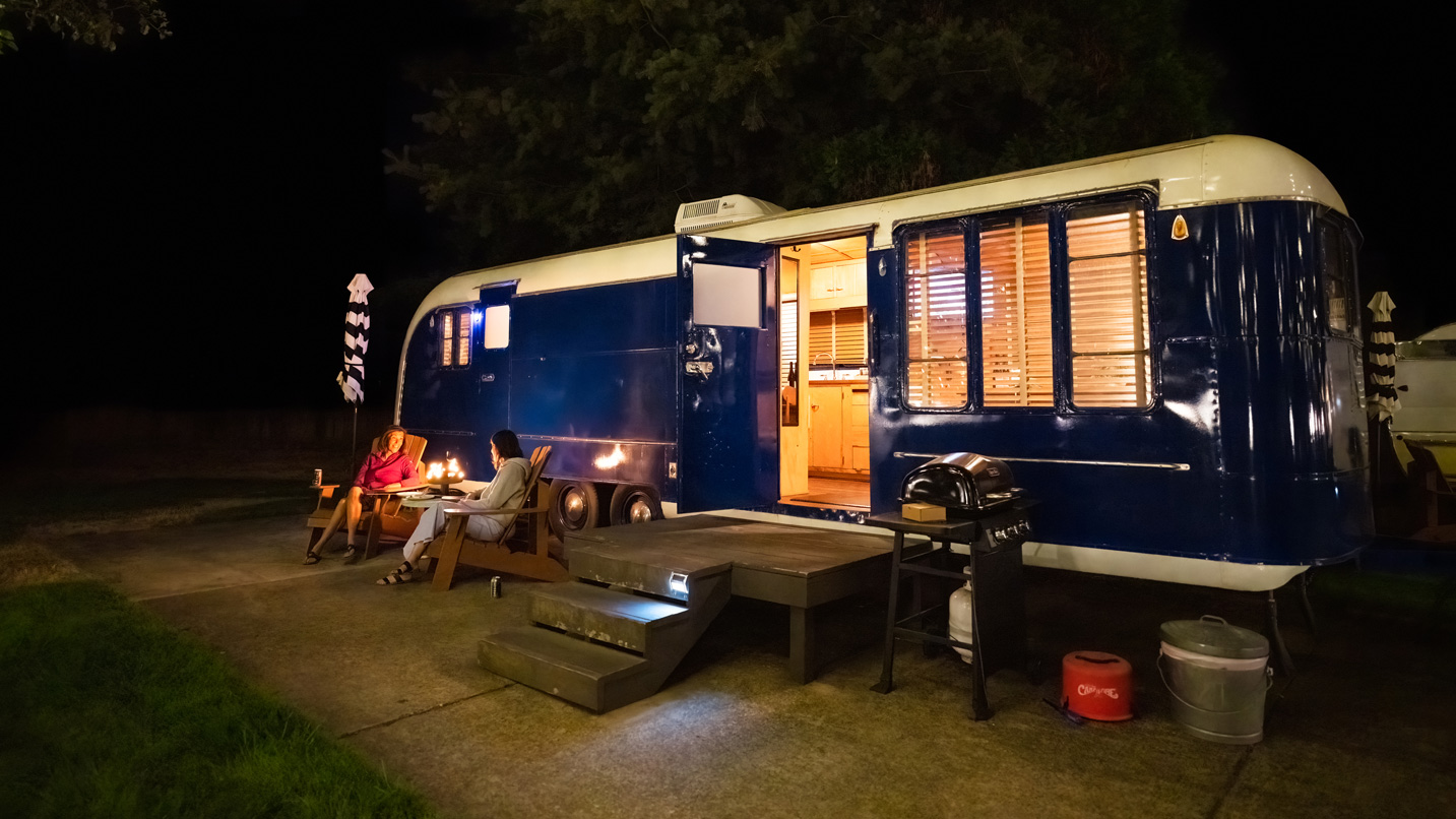 A retro-style RV camper at night. People sit outside near the camper at a small table.