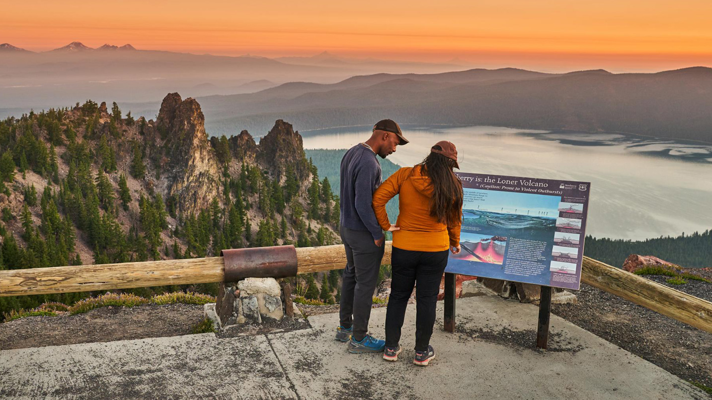 Two people look at an interpretive sign overlooking mountains and a lake.