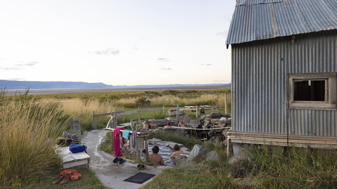 People in outdoor hot springs next to a sheet metal building.