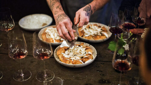 A chef puts together three dishes on a table surrounded by glasses of red wine.