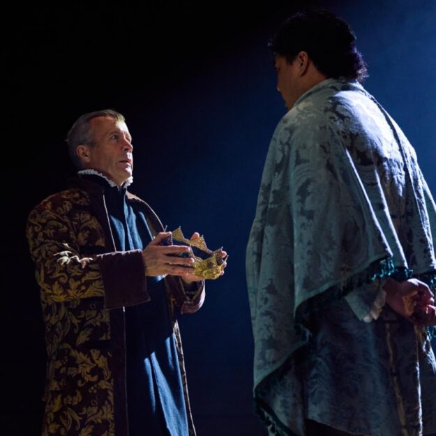 Two men wearing robes (one holding a crown) perform a theater scene against a dark blue background