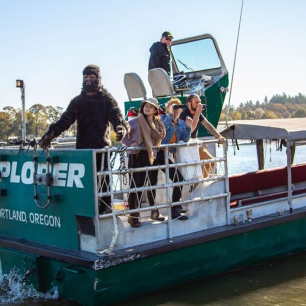 Four people stand on a small boat on the river next to a person in a Bigfoot costume