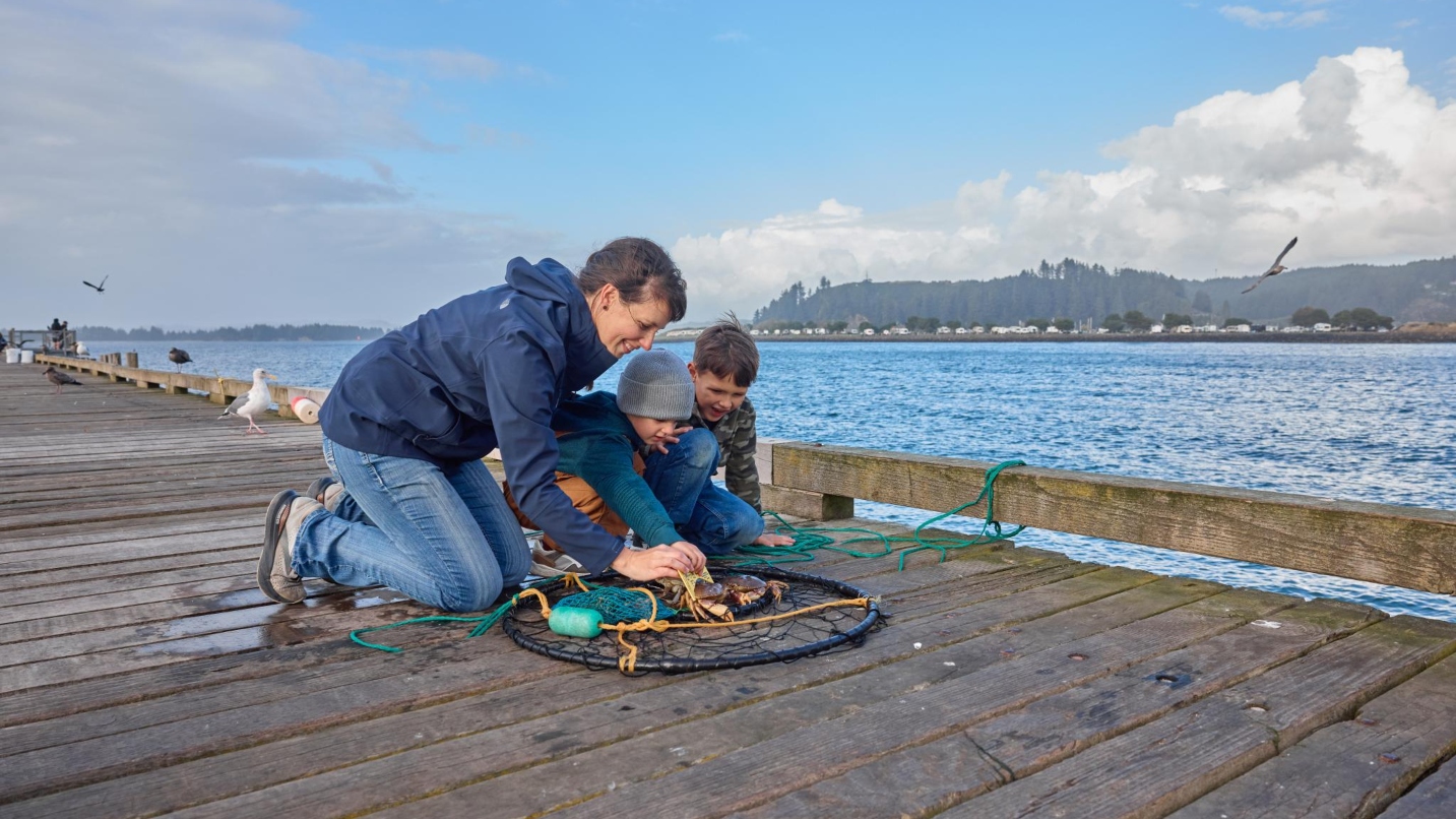 A woman and two young boys kneel on a wooden dock, measuring a fresh crab in a net with birds and blue sky in the background