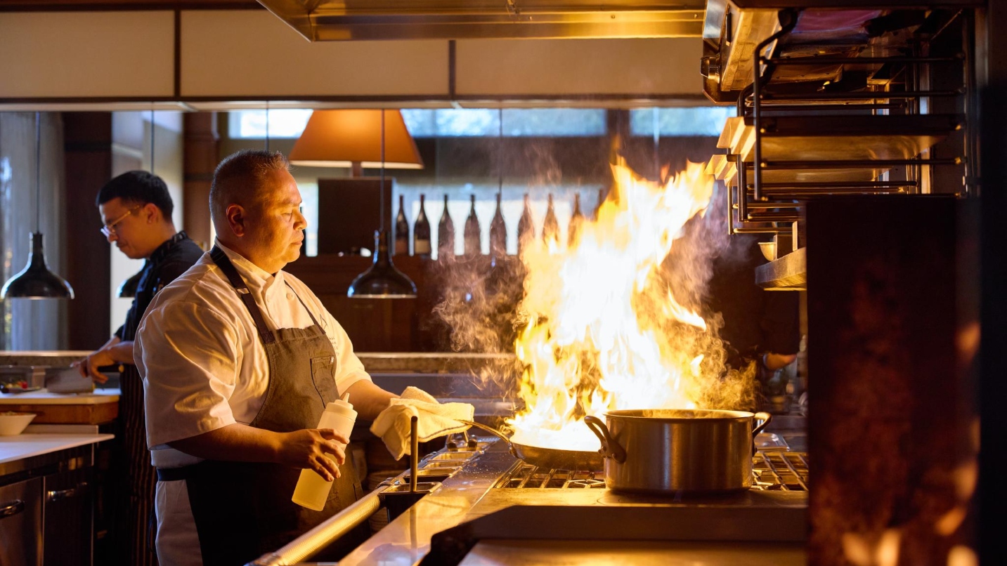 A chef wears an apron and holds the a bottle of oil and the handle of a flaming pan in a restaurant