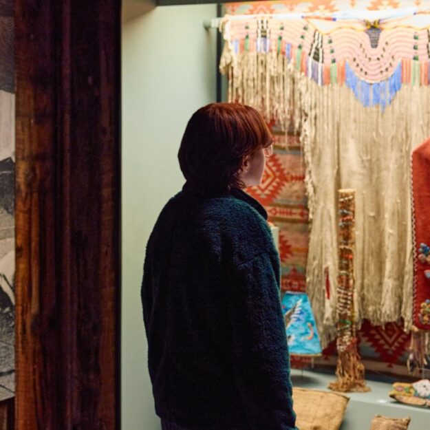 A person with short red hair looks at an exhibit of colorful Indigenous artifacts behind glass
