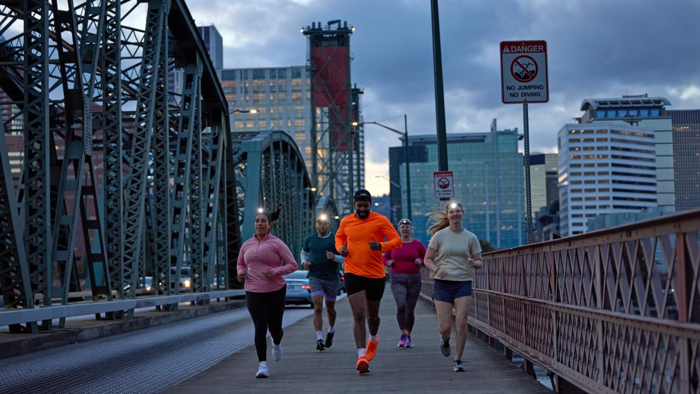 Five people wear headlamps and running clothes as they run across a bridge with a cityscape in background.