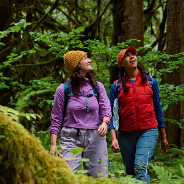 Two women in bright outdoor clothing and hats walk on a trail in a forest with moss and ferns in foreground