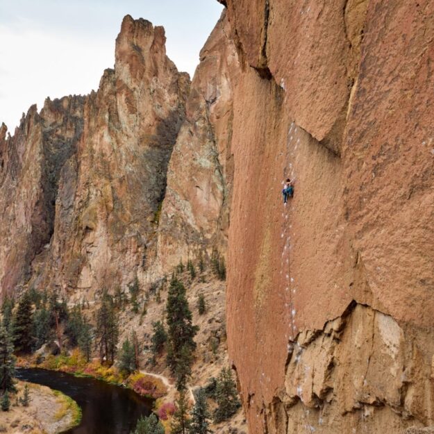 A rock climber climbs a big red rock wall in the distance with a river and trees below, and blue sky background
