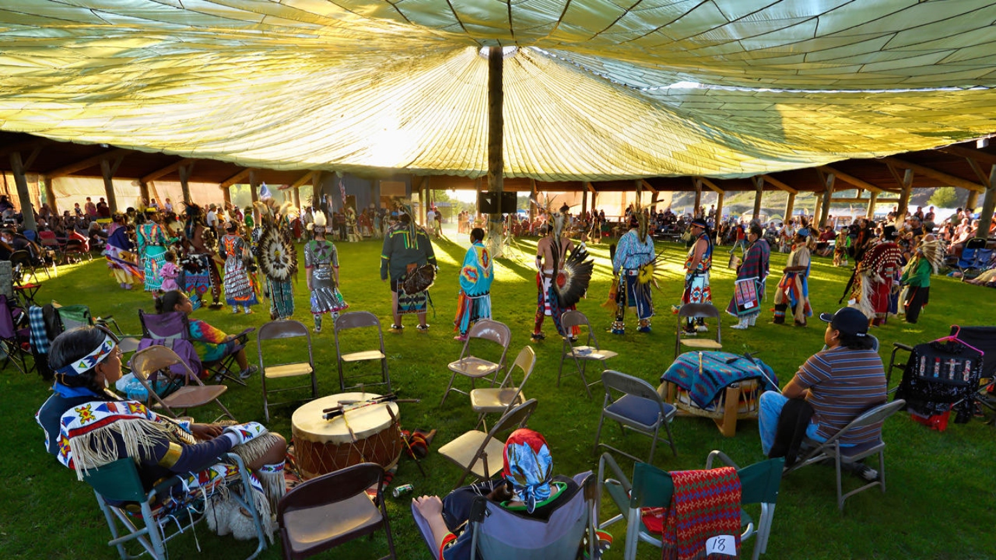 People stand under a tent in native regalia in a circle, with a few onlookers sitting in chairs outside the circle