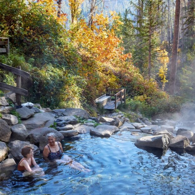 Two women sit in hot spring surrounded by rocks, a wooden boardwalk and trees of different colors