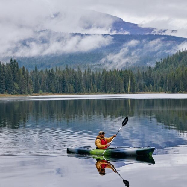 A man wears a lifejacket and holds a paddle while sitting in a kayak on a lake with mountains, trees and cloudy skies in the background