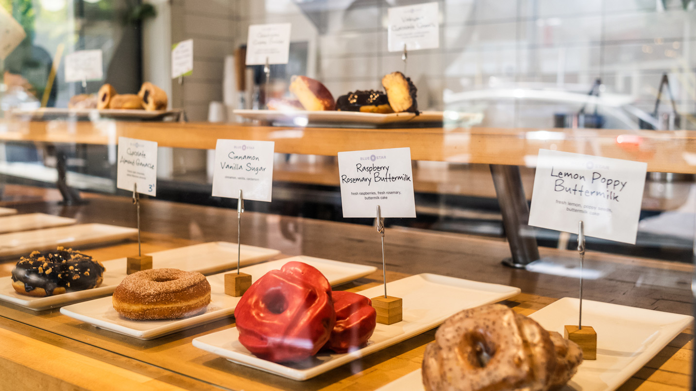 A display case of a variety of donuts, with signs on each variety stating the flavor. Some of the signs read Lemon Poppy Buttermilk, Raspberry Rosemary Buttermilk and Cinnamon Vanilla Sugar.