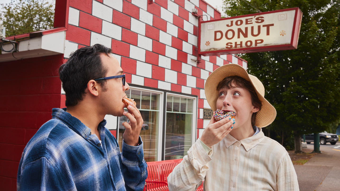 Two people bite into donuts while standing outside a red and white brick building, with a sign reading Joes' Donut Shop