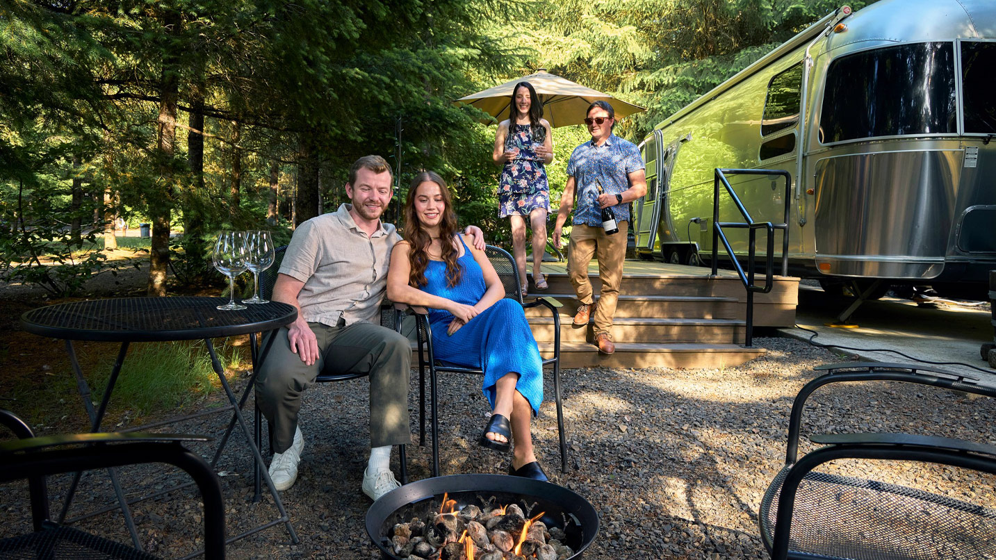 A couple sit on a gravel patio of an Airstream RV looking at small fire pit. Another couple in the background are walking towards the seating area.
