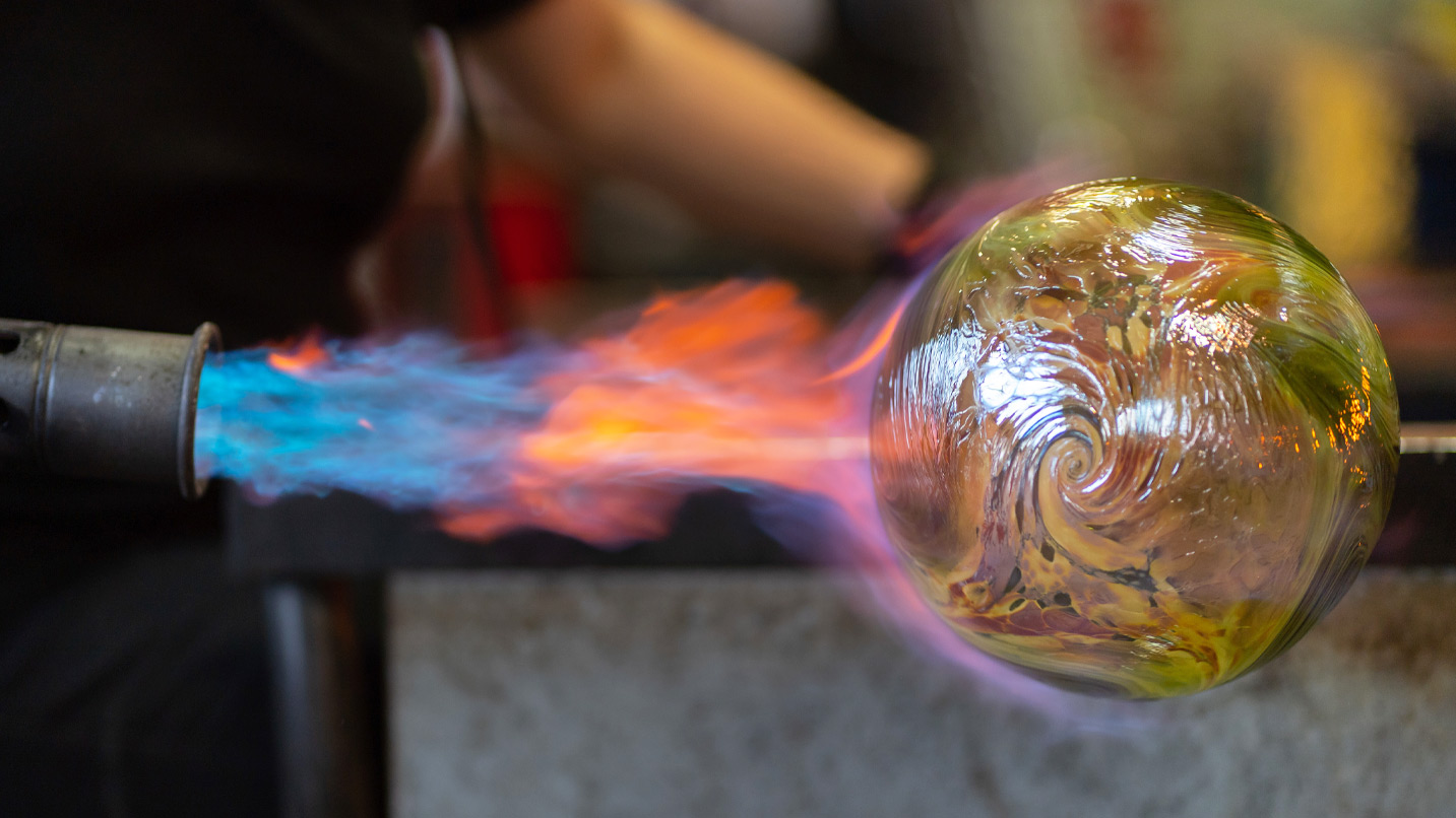 Closeup of a blowtorch firing onto a green glass rob with swirly design.