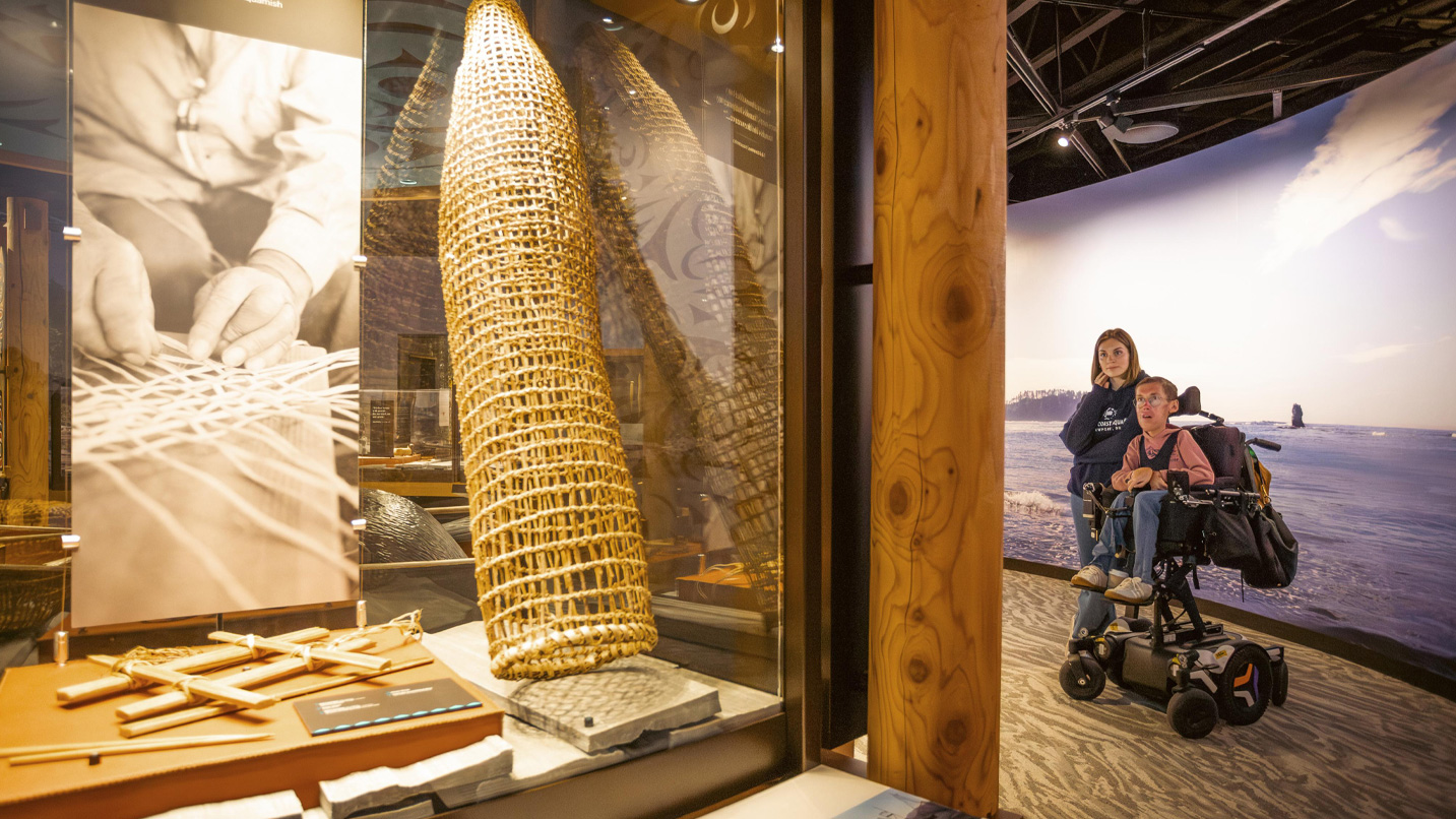 A couple, a woman and a man in a specialized, motor wheelchair, looking at a large weaved basket exhibit.