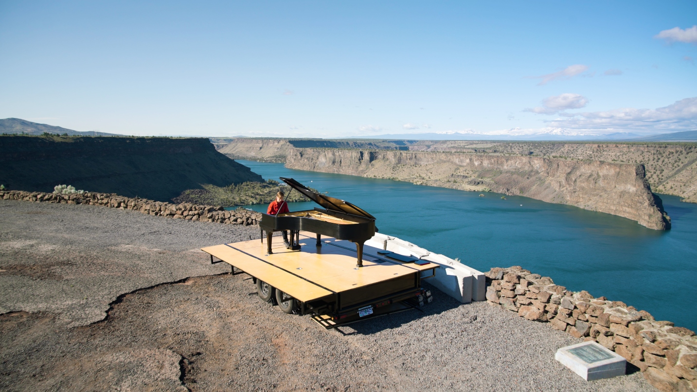 Man plays a piano on a makeshift stage next to a large lake with blue sky overhead