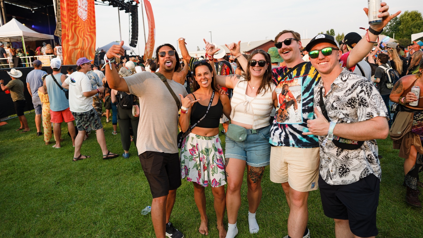 A group of five men and women wearing shorts, T-shirts and sunglasses smile and make hang loose signs at the camera, with a crowd of people behind them on the grass