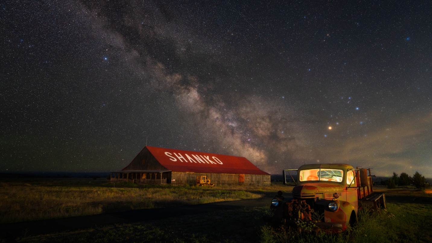 Starry night against a long building with the word Shaniko on the red roof, and an old yellow pickup truck in foreground