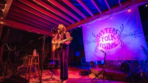 Man with long hair and beard plays guitar on stage with Sisters Folk Festival sign and purple spotlight in background