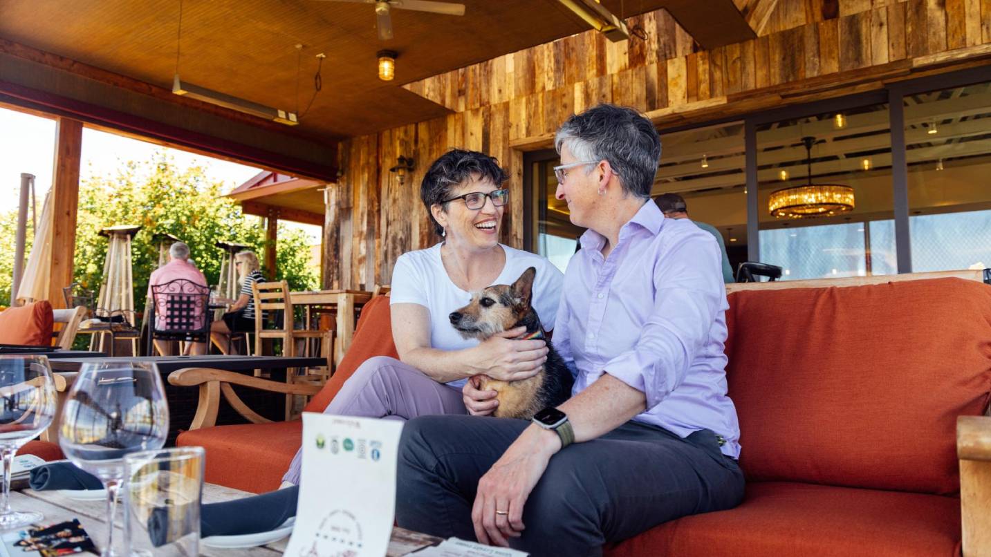 Two women on an outdoor patio couch with a small dog.