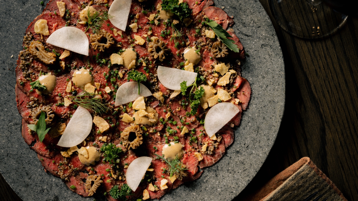 Modern grey stone round plate with thin-sliced beef carpaccio and various toppings, on a dark-wood table