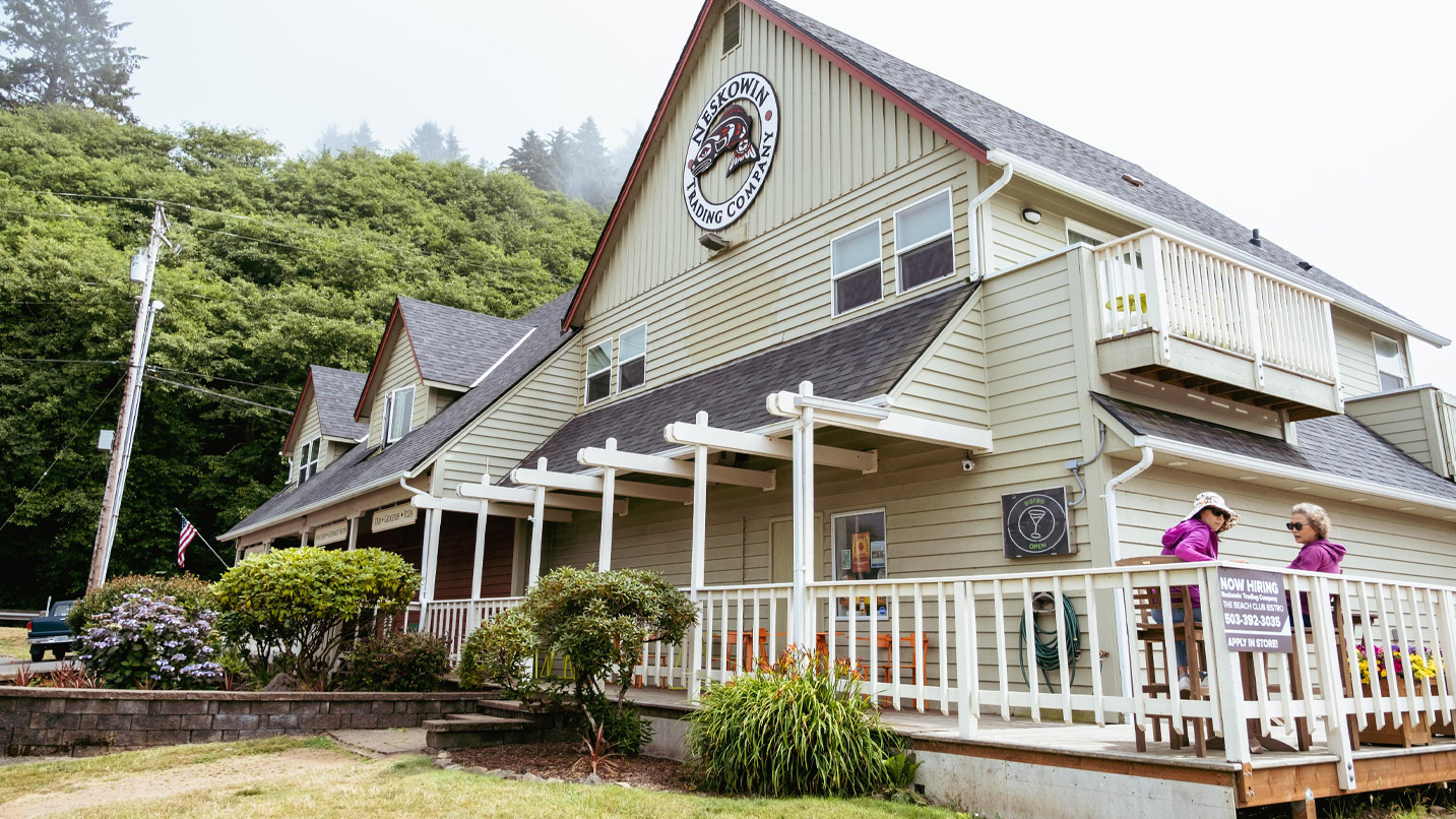 Exterior of commercial businesses building that look like a house, with a wrap around porch for seating. A large sign on a tall roof reads "Neskowin Trading Company."