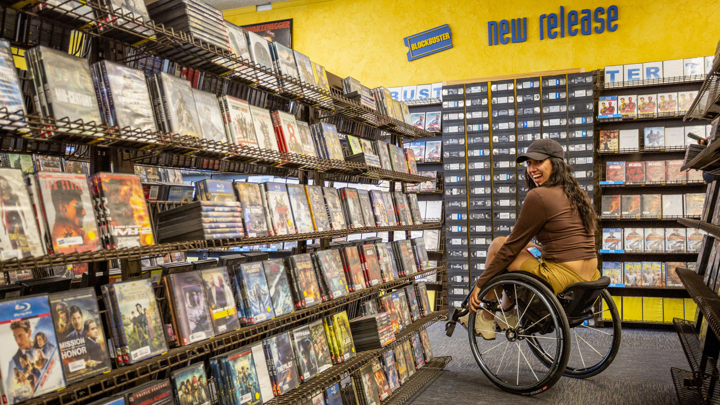 A smiling woman in a wheelcair amongst shelves of DVDs. On the wall a small Blockbuster logo sign and lettering reads "New Release" under a shelf of DVDs.