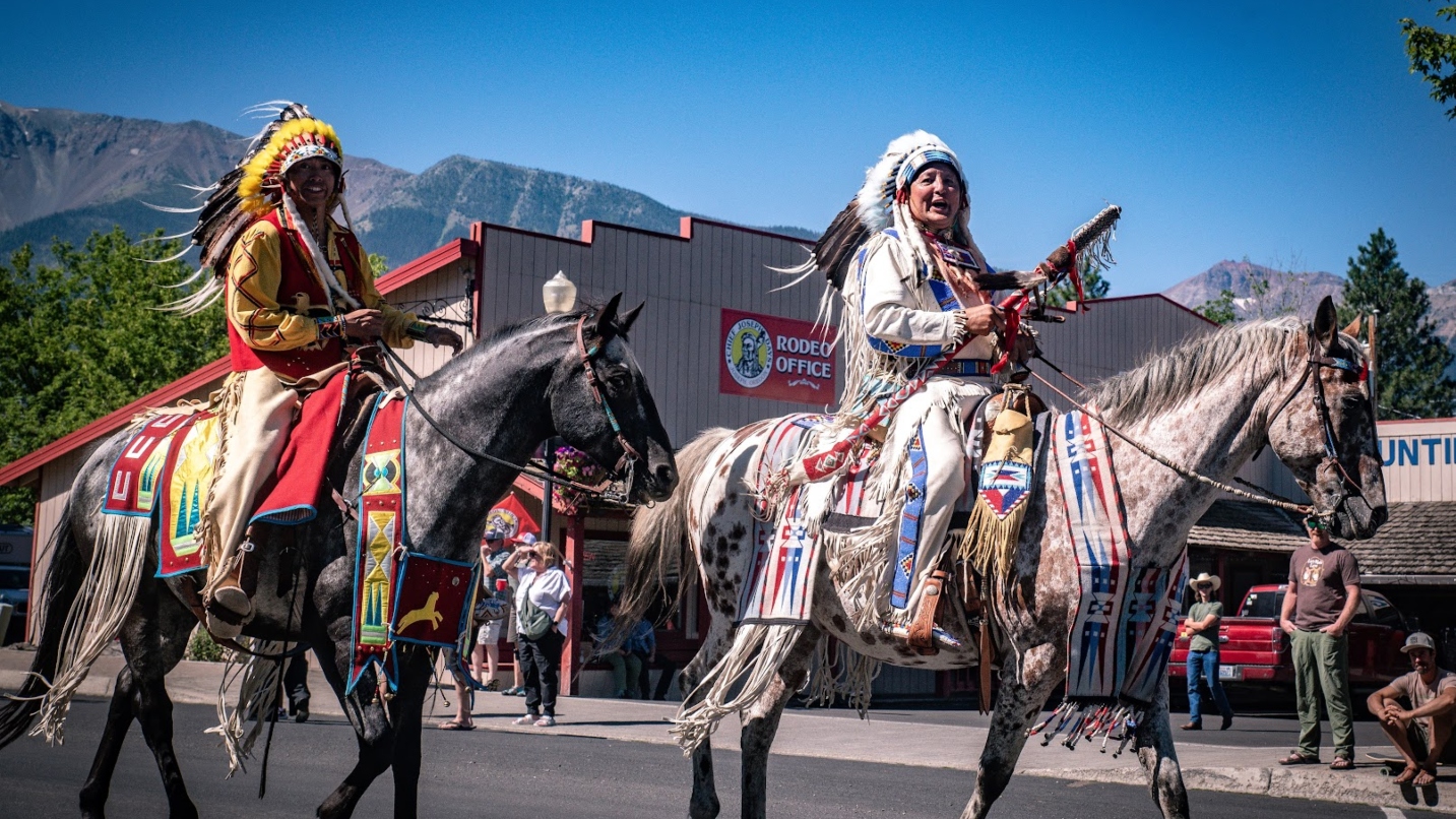 Two men wear Native regalia and ride on horseback in a parade with a building, mountain range and blue sky in the background.