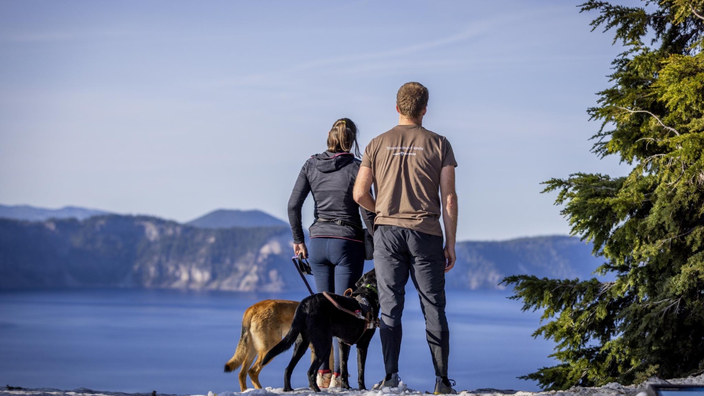 Man, woman and two dogs on leash look out on lake in a caldera with fir tree in foreground and blue sky