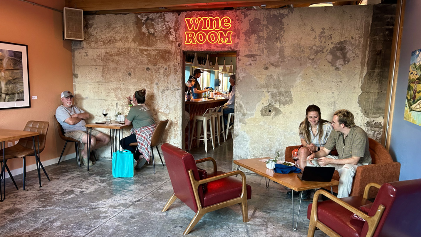 People sitting at tables inside a small lobby with food and drinks. A doorway to a bar area has a neon sign overhead that reads "Wine Room"