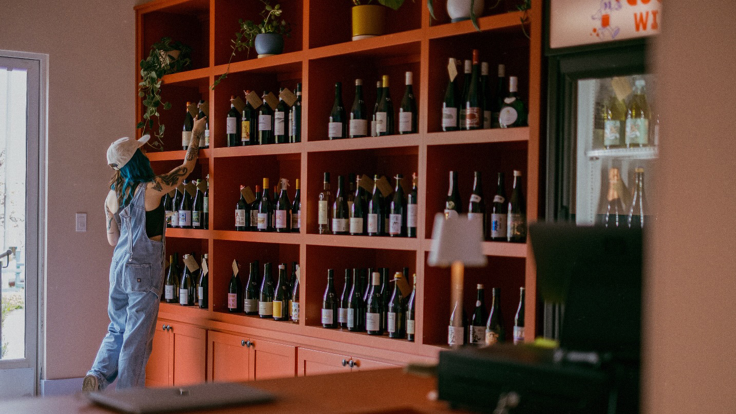 A woman reaching for a bottle of wine from a large shelf of bottles.