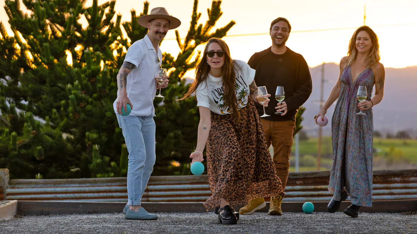 Four friends at sunset drinking wine and rolling bocce balls.