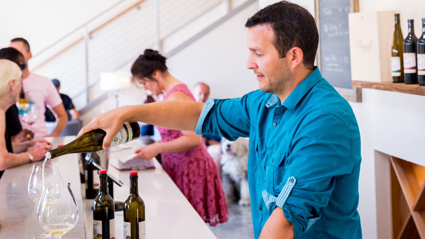 A man behind a counter pours wine into a glass.