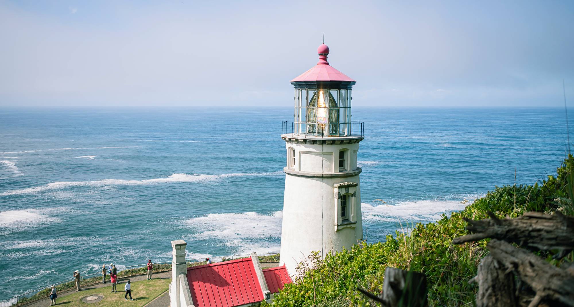 Un faro con la parte superior roja, situado en una colina con una vista que se extiende hasta el océano. Hay personas tomando fotos.