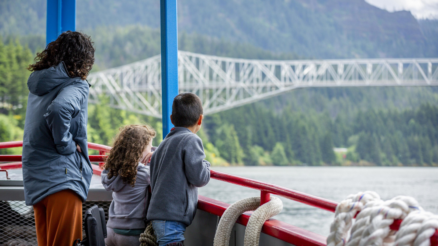 An adult woman and two young children look from aboard a boat towards the Bridge of the Gods.