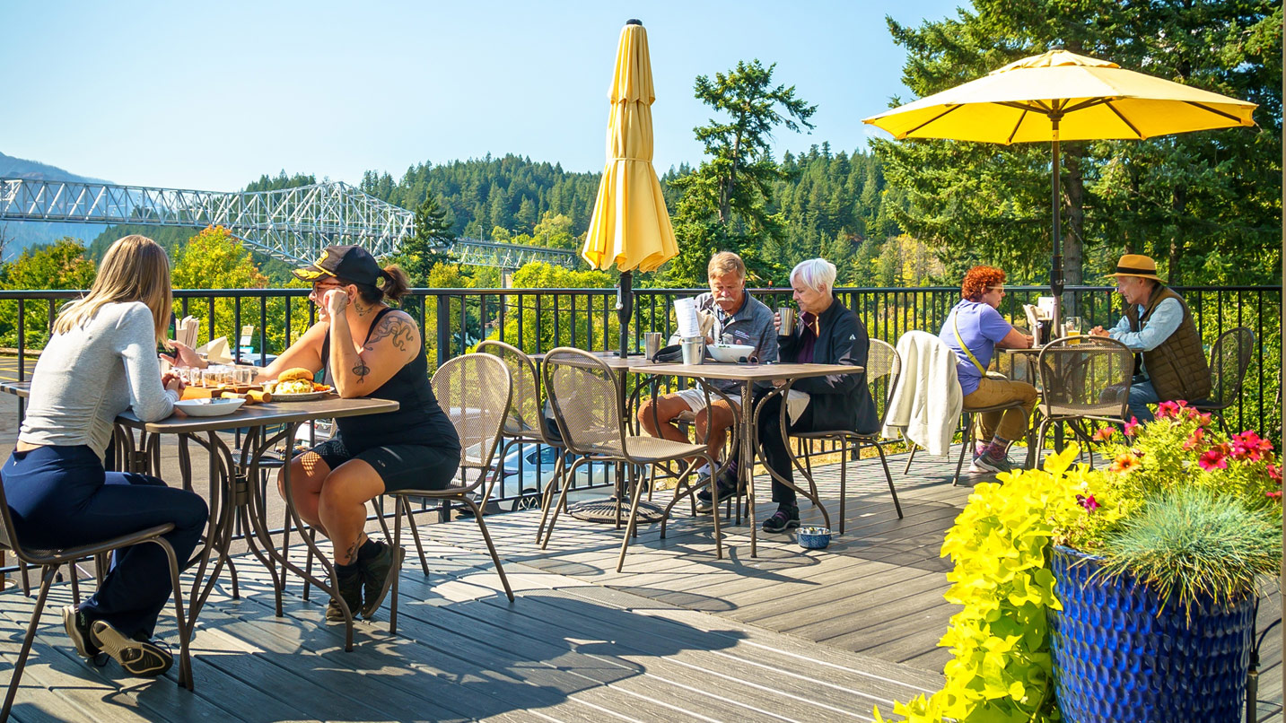 Several people sit at an outdoor patio on a sunny day. The Bridge of the Gods is visible from the patio.