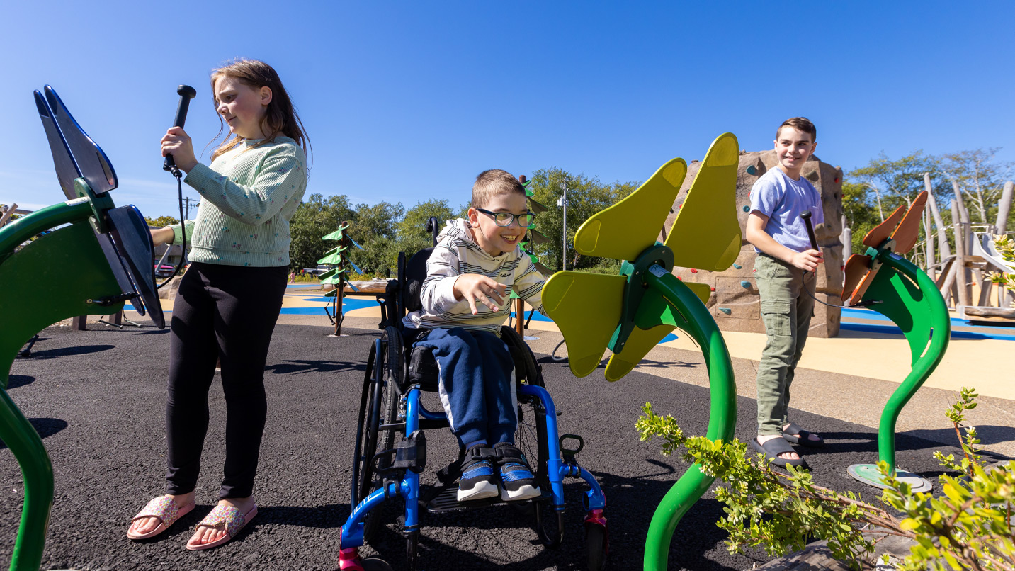 Three children, one in a wheelchair, play with accessible play structures.