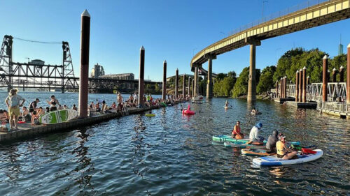 Several people swimming, paddleboarding and sitting on a long dock on the Willamette River. In the background, the Steel Bridge and part of the Portland skyline are visible.